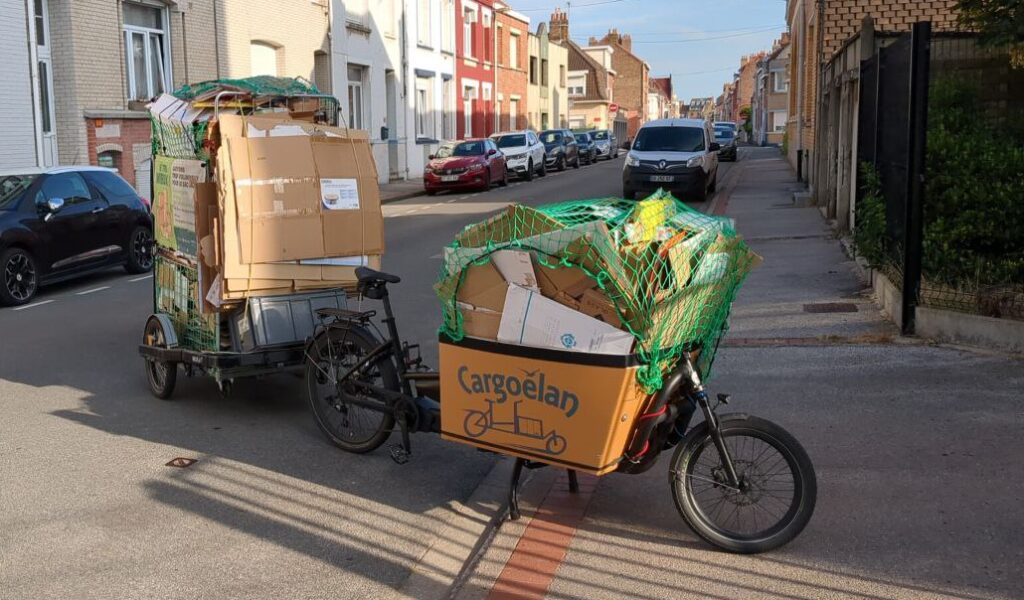 un vélo cargo rempli de cartons sur l avant et sa remorque pleine de cartons en vue de profil dans les rues de malo les bains .