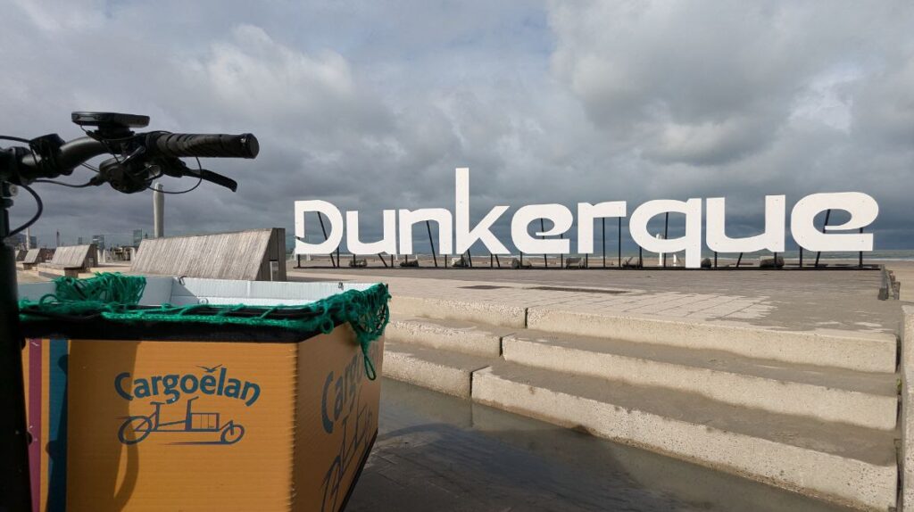 à gauche de l'image une partie de vélo cargo sur lequel il y a le logo cargoélan placé face à un lettrage "Dunkerque" disposé sur la digue de Malo les bains avec en arrière plan la plage et la mer sous un ciel de nuages bas.